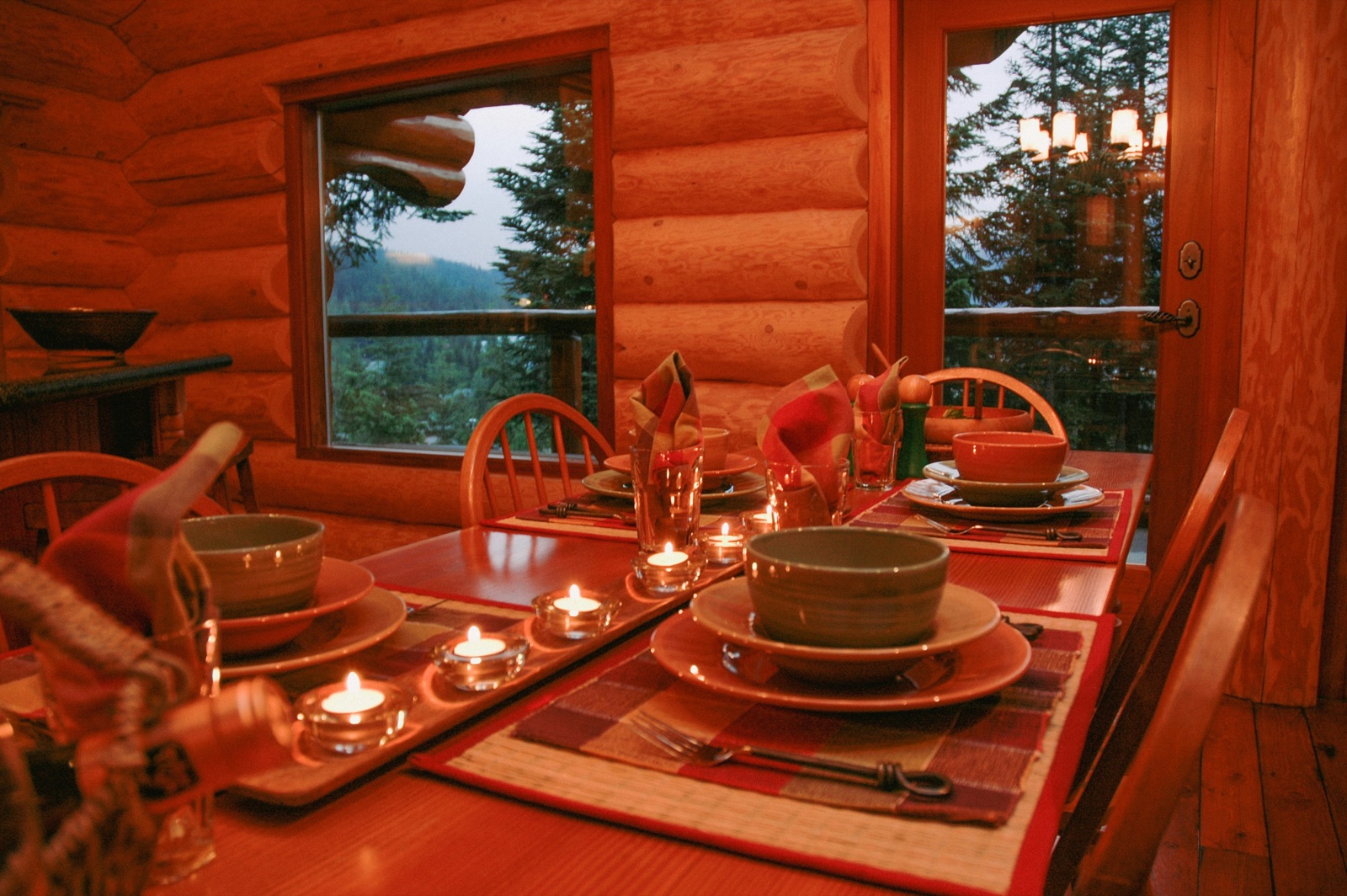 A candlelit table set for a gathering inside a log cabin, with evergreens and mountains through the windows